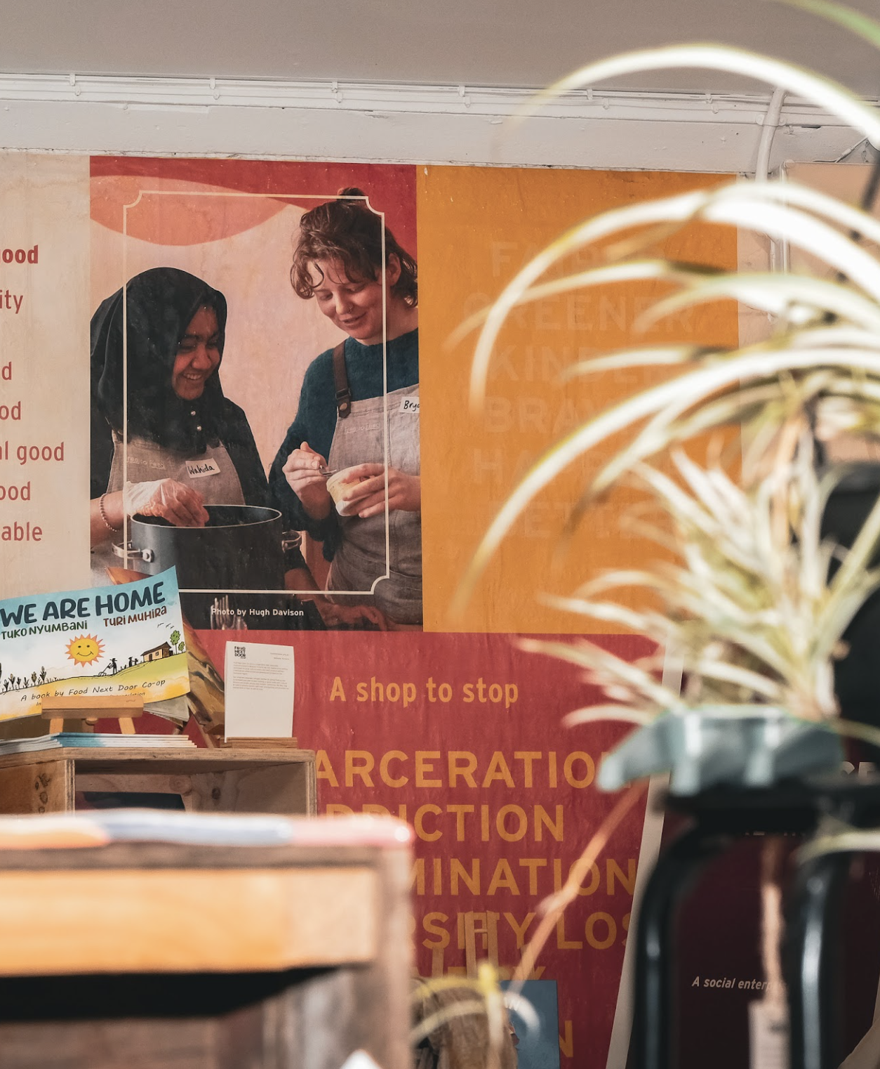 A welcoming social enterprise retail space featuring books, plants and social impact displays, with a large photograph of two people preparing food together that reflects community connection, participation and collaboration.